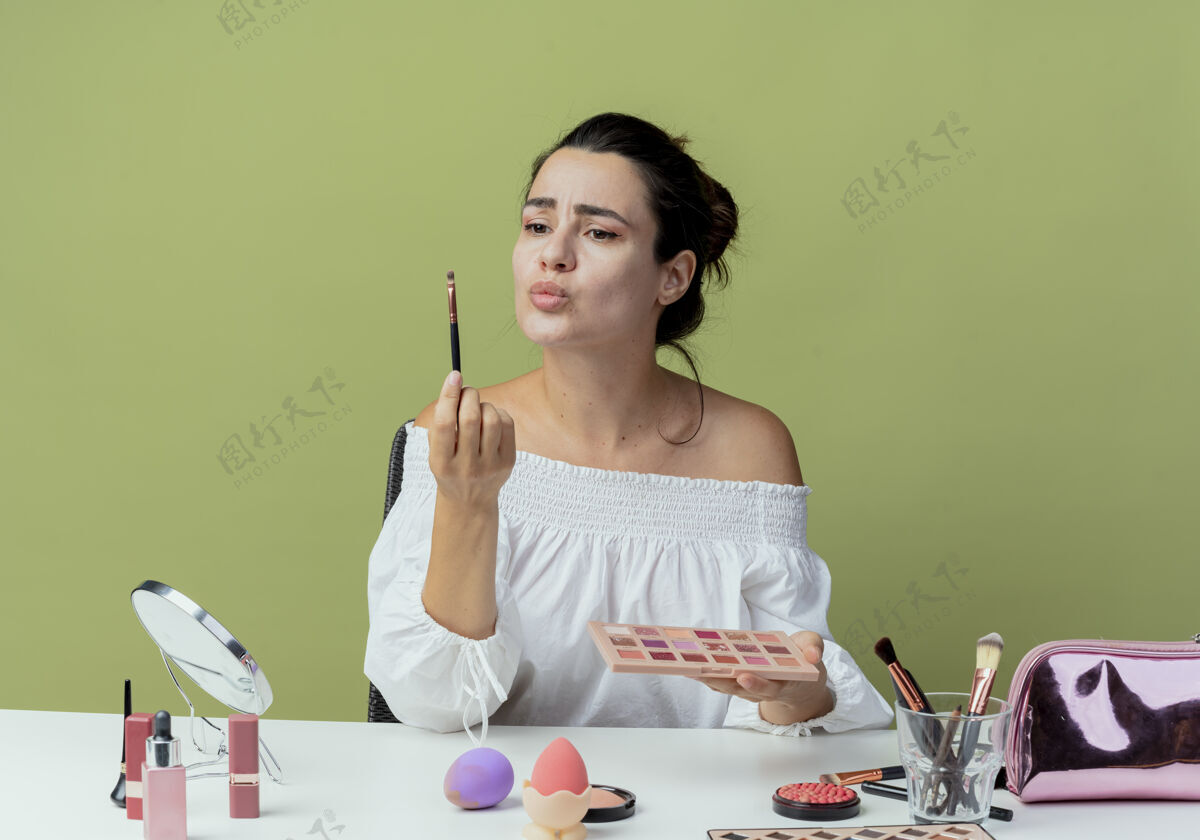 坐迷茫的美女坐在桌上 闭着眼睛 用化妆工具拿着眼影调色板和化妆刷 隔在绿色的墙上拿着桌子迷惑