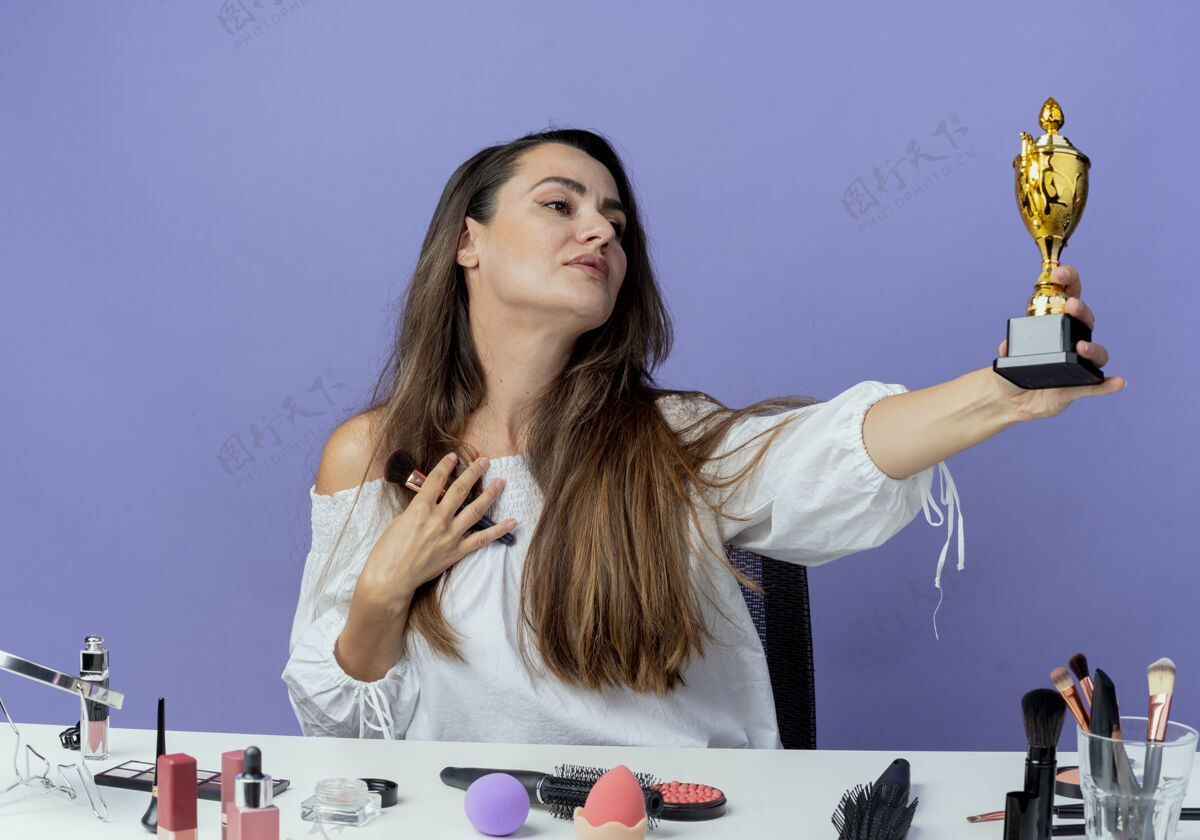 拿着高兴的漂亮女孩坐在桌旁 拿着化妆工具 拿着化妆刷 看着紫色墙上孤立的优胜者杯请坐优胜者美女