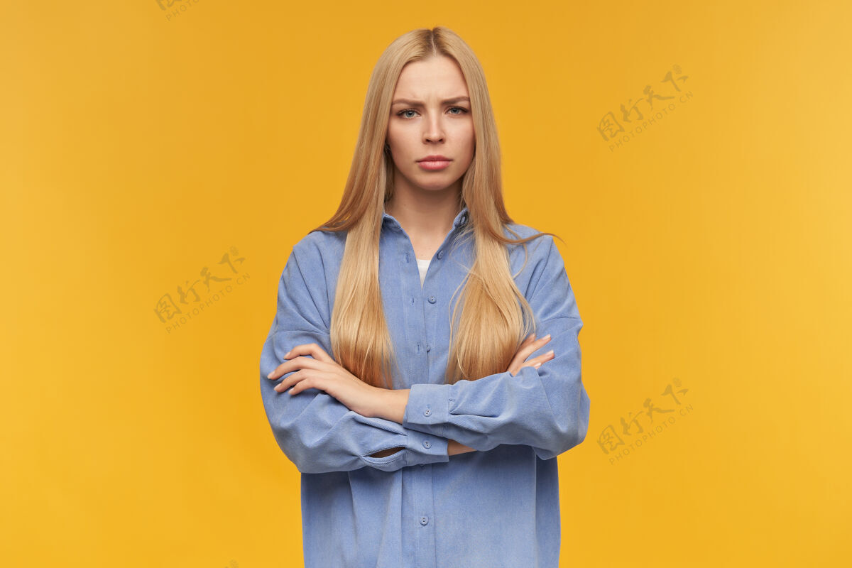 女孩严肃的女人 美丽的女孩 金色长发穿着蓝色衬衫人和情感的概念保持双臂交叉在胸前看着镜头 橙色背景下孤立衬衫女性20多岁