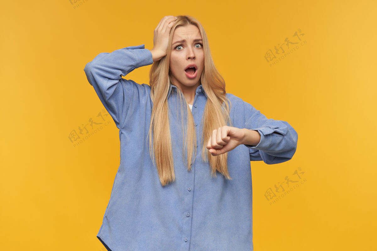 晚有压力的金发女孩的肖像穿着蓝色衬衫人和情感概念触摸她的头 检查手表 时间看着相机 隔离在橙色背景上服装20多岁女士