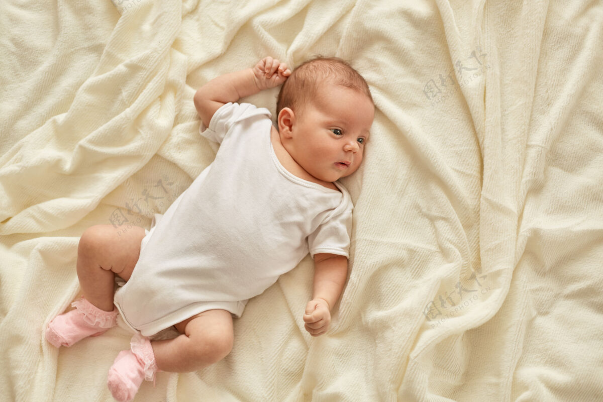 女孩刚出生的女婴或男婴躺在床上的毛毯上望着别处 穿着白色的紧身衣和袜子 婴儿在学习周围的世界 有着困倦的表情新的毯子婴儿