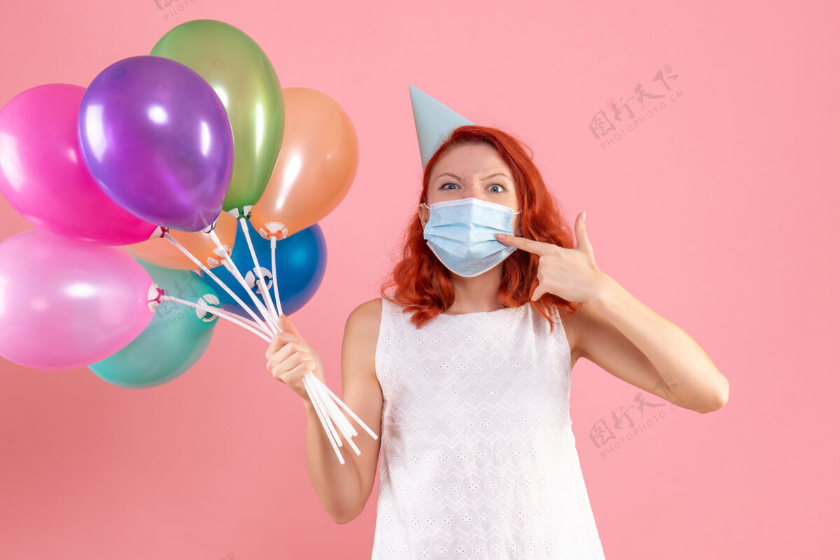 气球正面图：年轻女性戴着面具手持彩色气球站在粉色办公桌上圣诞彩色病毒大流行-派对举行病毒圣诞节