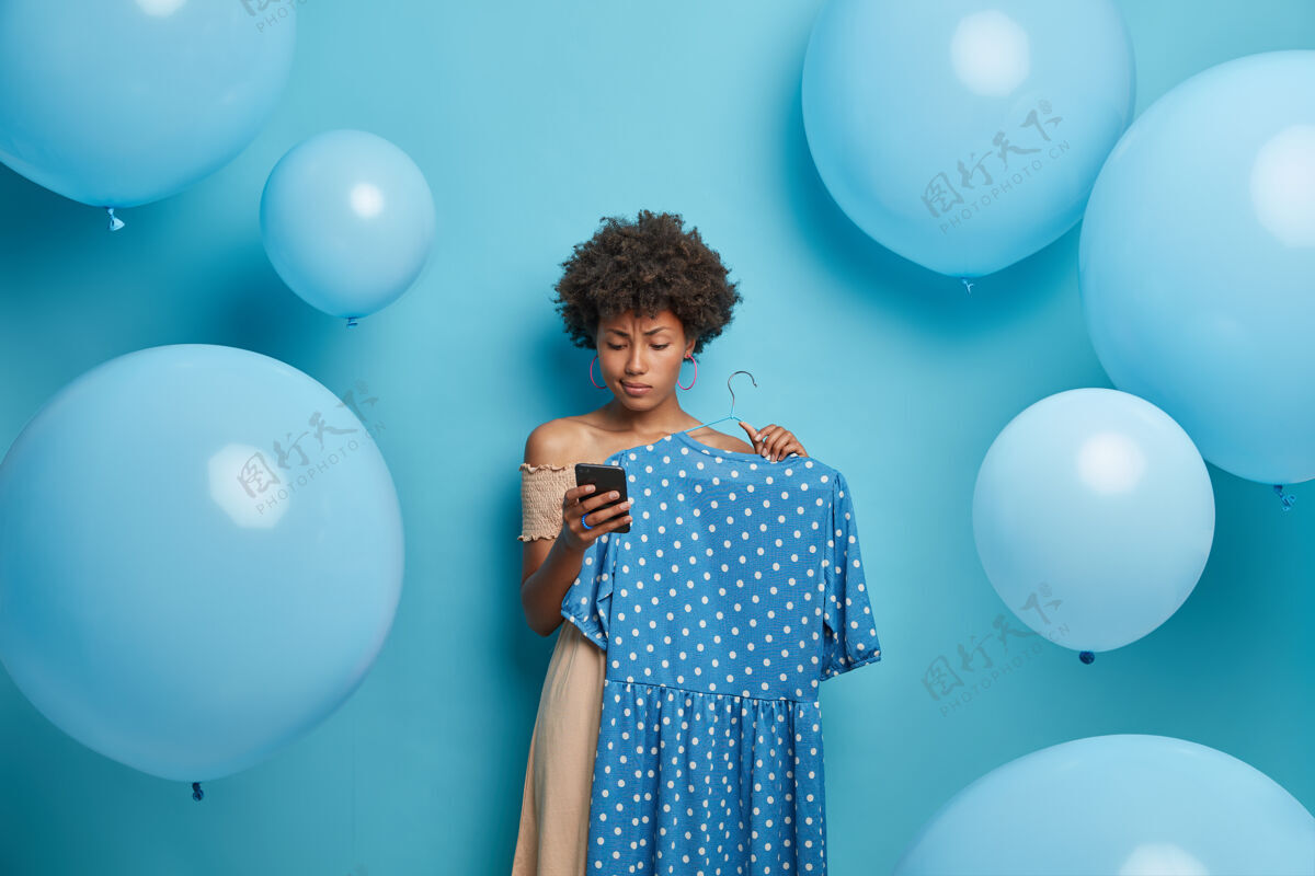 服装严肃的生日女孩在智能手机上收到祝贺 在衣架上挑选蓝色圆点连衣裙 穿好衣服等待客人 站在装饰好的墙上女人 衣服 穿衣不高兴优雅女人