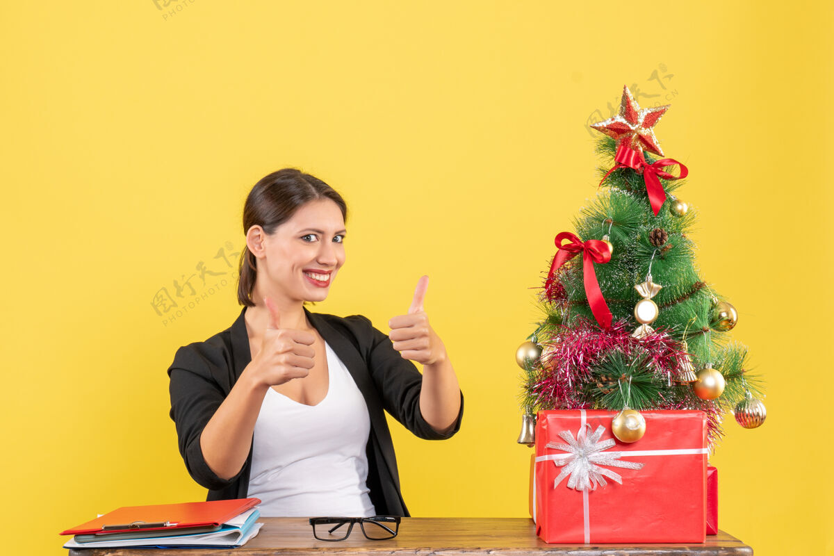 制造在黄色的办公室里 身着西装的年轻女子微笑着在装饰好的圣诞树旁做着“ok”的手势手势靠近微笑