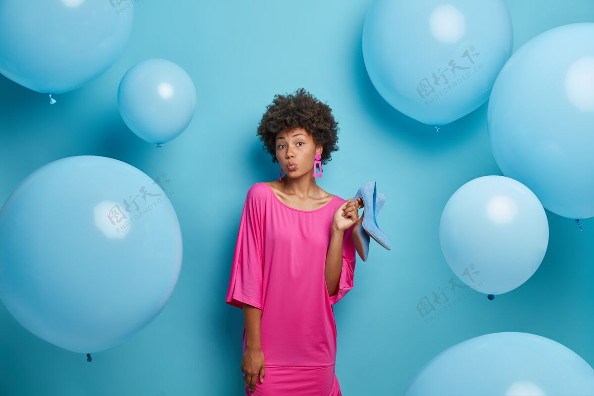 衣柜可爱的黑皮肤卷曲的非洲裔美国女人保持嘴唇圆润 穿着时髦的衣服 拿着鞋子 选择穿什么或穿什么 隔离在蓝色的墙上 周围有充气的气球穿着气球衣服
