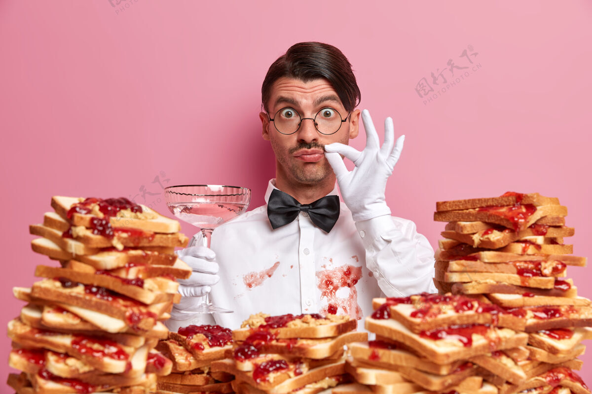 白种人专业服务员端着一杯含酒精的鸡尾酒站着 展示着完美的味觉标志 在吃了美味的三明治后 穿着被果酱弄脏的白衬衫 隔离在粉红色的墙上服务和餐饮手套职业惊喜