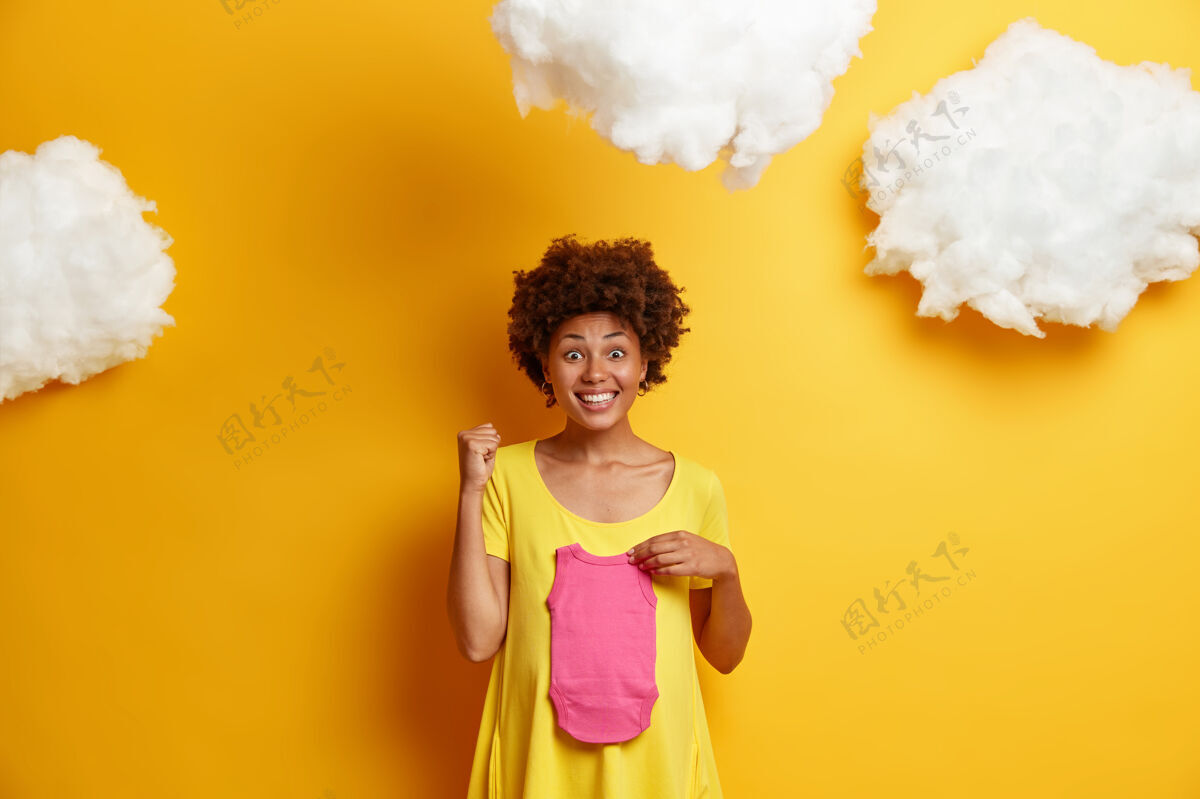 服装快乐的非洲裔美国妇女握紧拳头 发现自己将要生女儿时感到高兴 把婴儿抱在肚子上 顶着黄色的云朵站在上面怀孕的概念腹部等待请