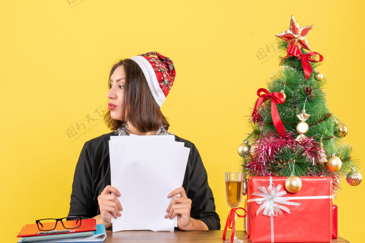 圣诞老人穿着西装 戴着圣诞老人帽和新年装饰品 手里拿着文件 坐在办公桌旁 桌上放着圣诞树坐着年文件