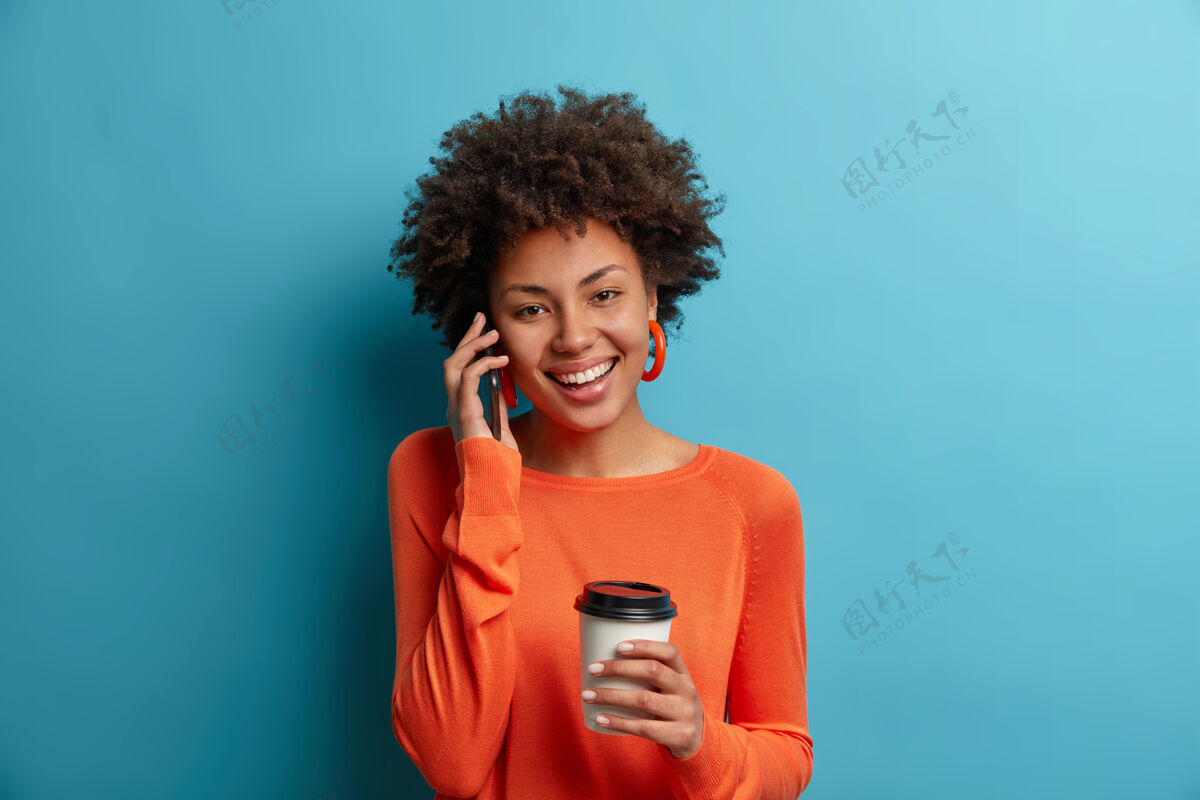 室内快乐无忧无虑的千禧一代少数民族女孩有愉快的电话交谈 把智能手机放在耳边 有可爱的微笑和愉快的心情 喝着芳香的饮料 穿着橙色的套头衫 孤立在蓝色上请表达牙关