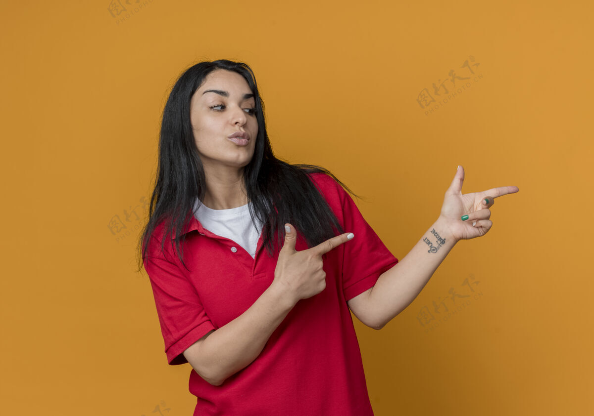 穿自信的年轻黑发白人女孩穿着红色衬衫 两手孤立地站在橙色的墙上 看着并指着旁边二黑发红色