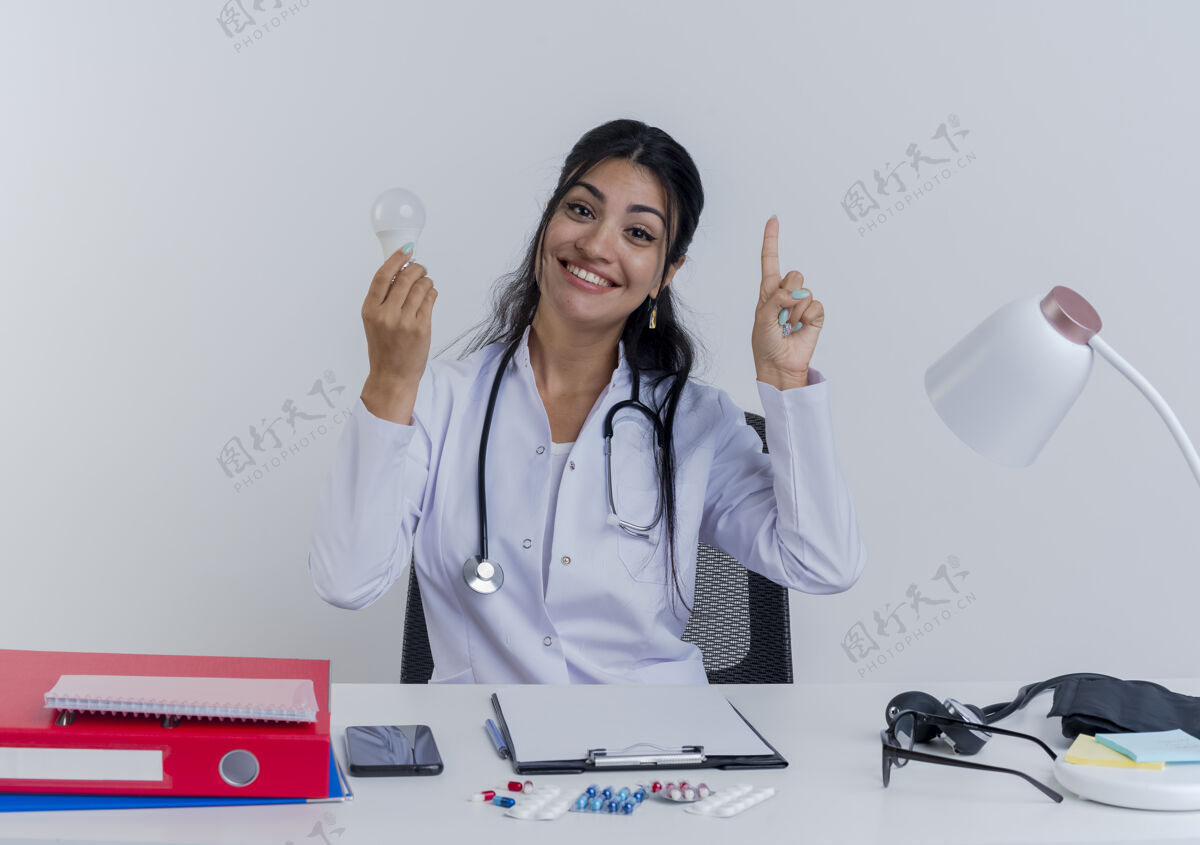 微笑面带微笑的年轻女医生穿着医用长袍和听诊器坐在办公桌旁 手里拿着医疗工具 手里拿着光泡 抬起手指工具女士穿上