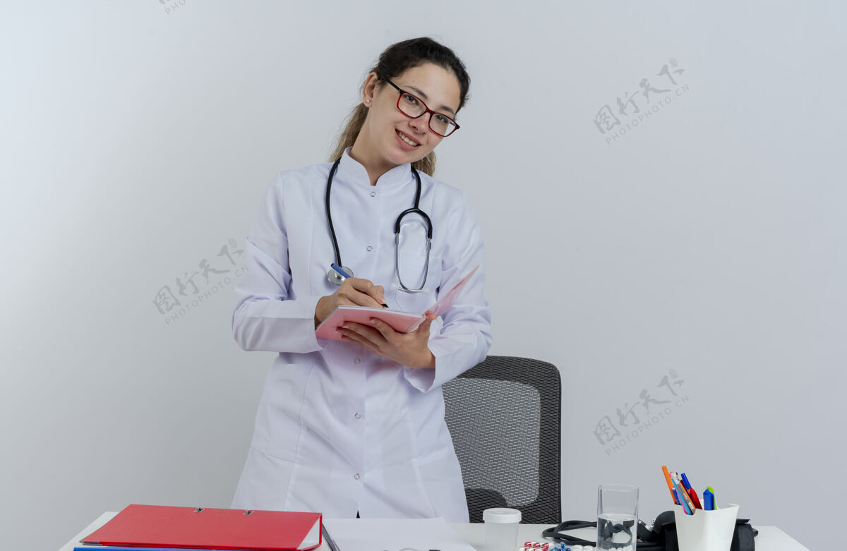 人面带微笑的年轻女医生 穿着医用长袍 戴着听诊器和眼镜 站在桌子后面 手里拿着医疗工具 看着用笔在笔记本上写字笔书写医生