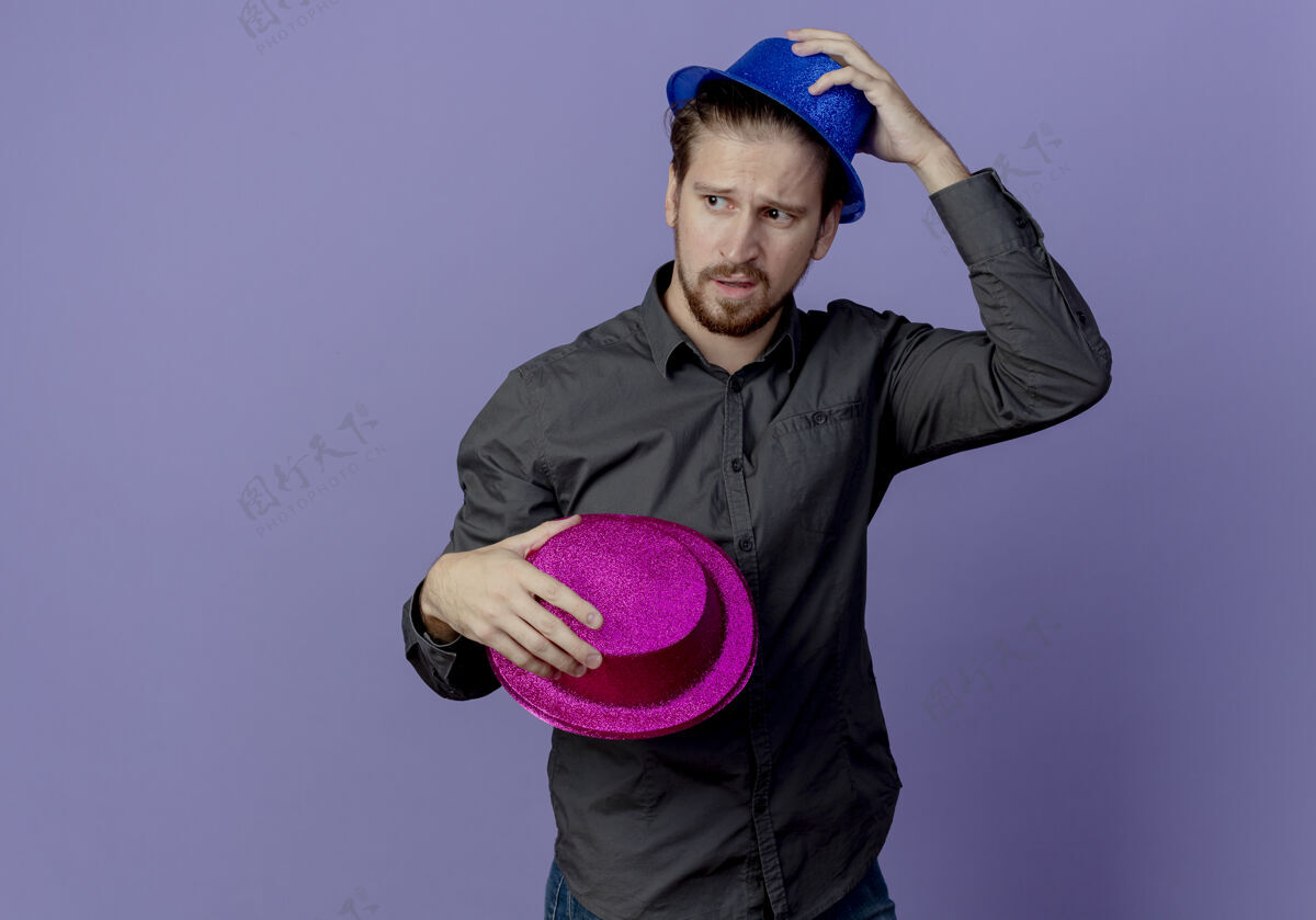 粉色恼羞成怒的帅哥拿着粉色的帽子 头上戴着蓝色的帽子 隔离在紫色的墙上帅气恼火男人