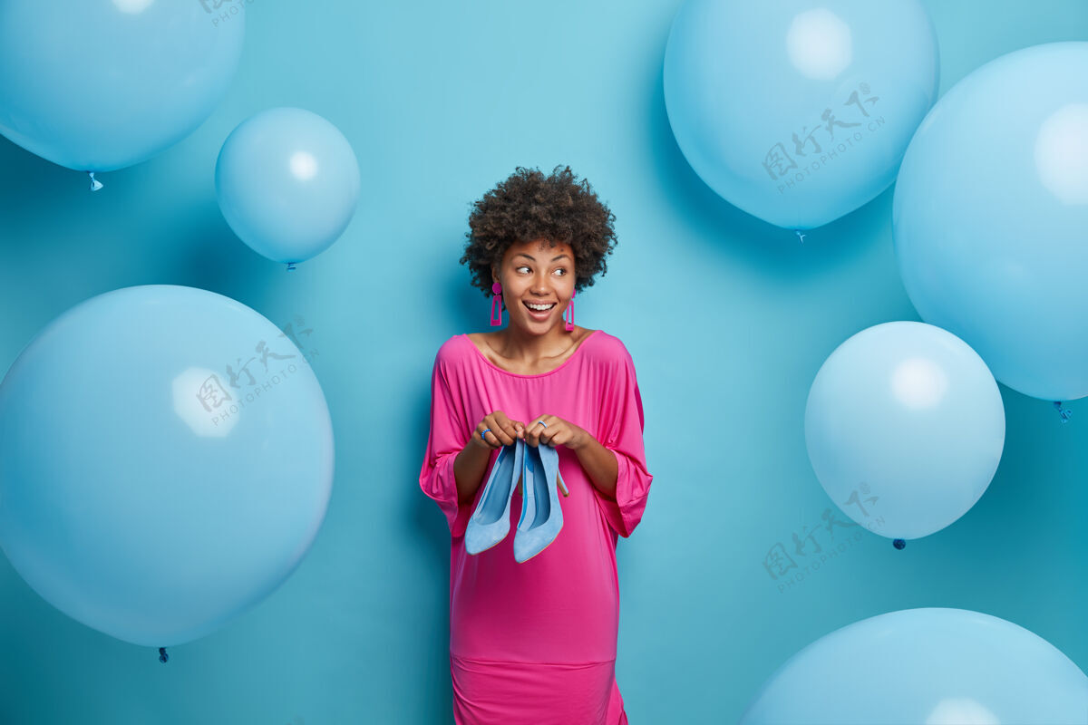 气球室内拍摄的是一个时髦的快乐女人 一头非洲头发 穿着粉红色的裙子 穿着高跟鞋 准备生日派对 试着选择穿什么 一边看 蓝色的气球在周围乐观节日年轻