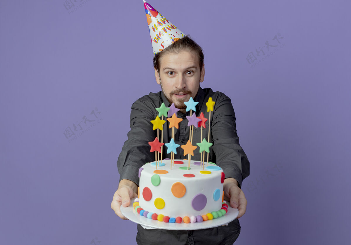 紫色戴着生日帽的快乐帅哥把生日蛋糕孤立地放在紫色的墙上男人帅哥蛋糕