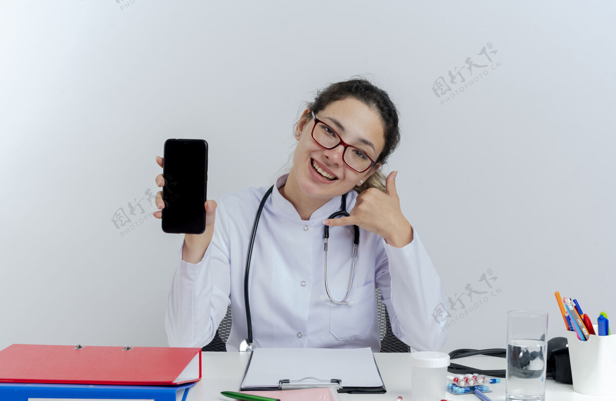 听诊器快乐的年轻女医生穿着医用长袍 戴着听诊器 戴着眼镜 坐在办公桌旁 拿着医疗工具 展示着手机 看起来在做通话手势欢乐手机医生