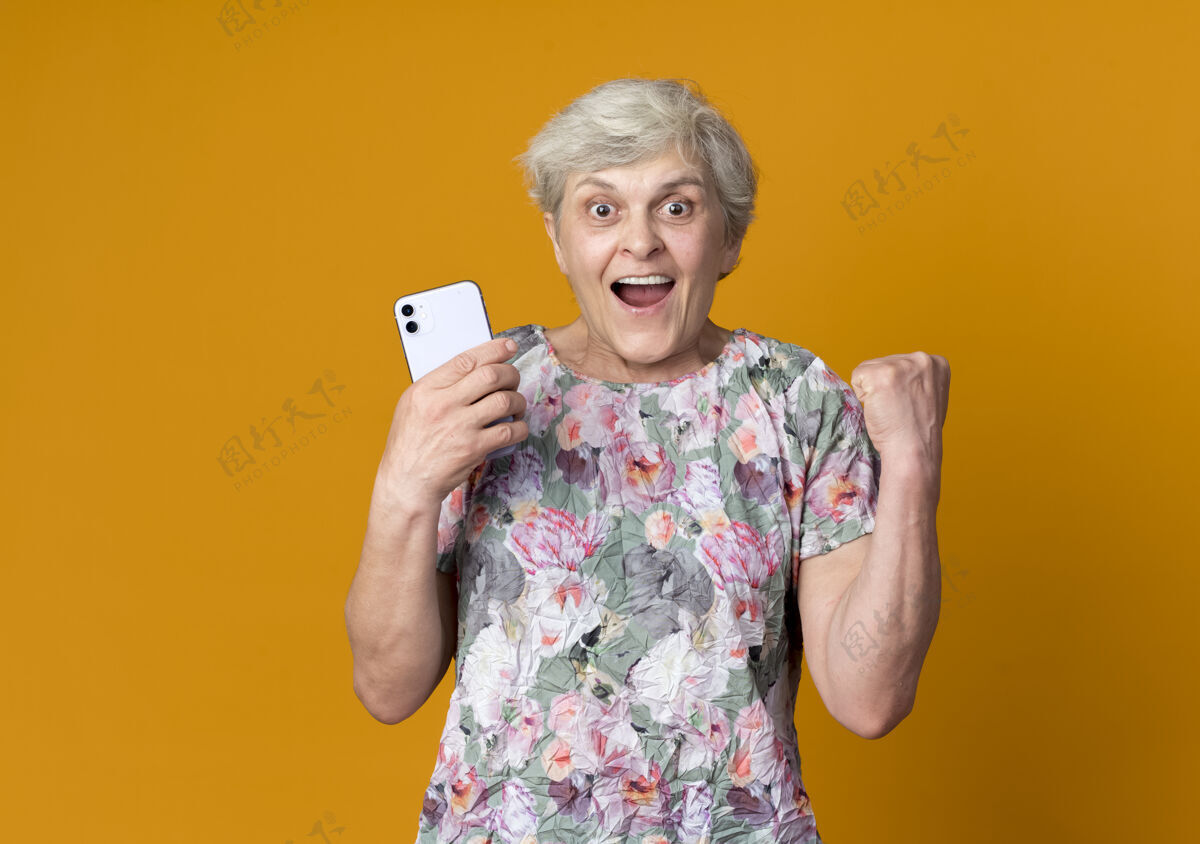 女人兴奋的老妇人举起拳头拿着电话孤立在橙色的墙上兴奋举起老人