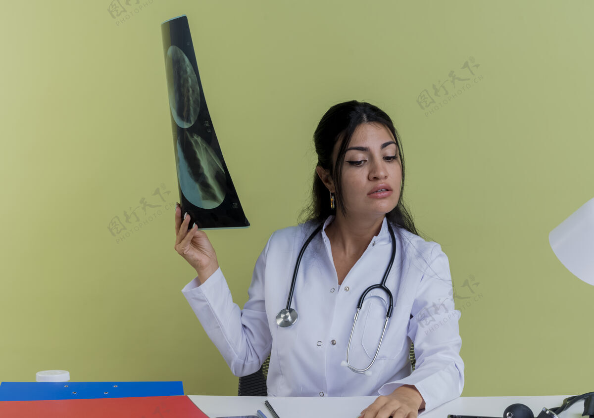 手自信的年轻女医生穿着医用长袍 手持听诊器坐在办公桌旁 手持医疗工具 手持x光片 手放在办公桌上 孤立地看着办公桌桌子成人自信