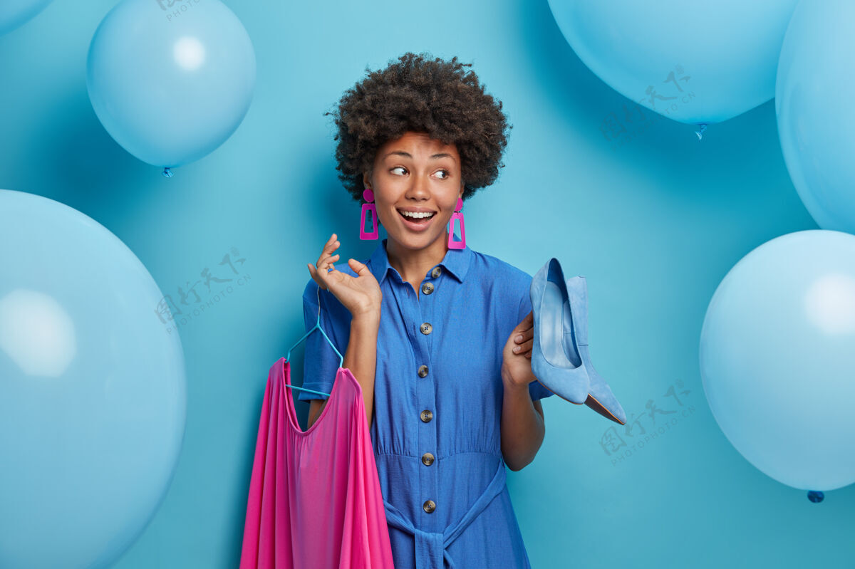 高兴高兴的积极的非裔美国女士选择外出约会 在衣架上拿着蓝色高跟鞋和玫瑰色连衣裙 准备参加聚会和庆祝活动 在蓝色的墙上摆着充气气球选择欢乐卷发
