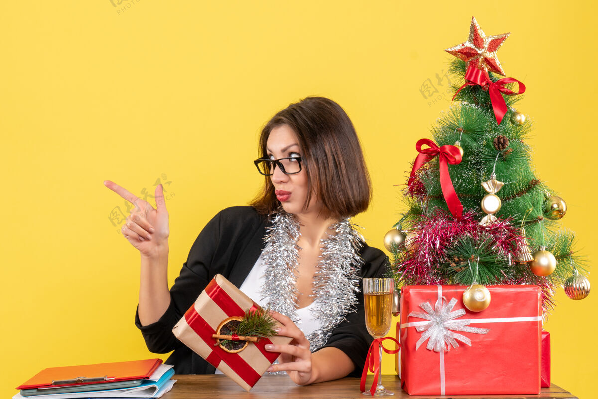 漂亮漂亮的商务女士 穿着西装 戴着眼镜 指着坐在办公桌旁的东西 桌子上有一棵圣诞树插花微笑什么的