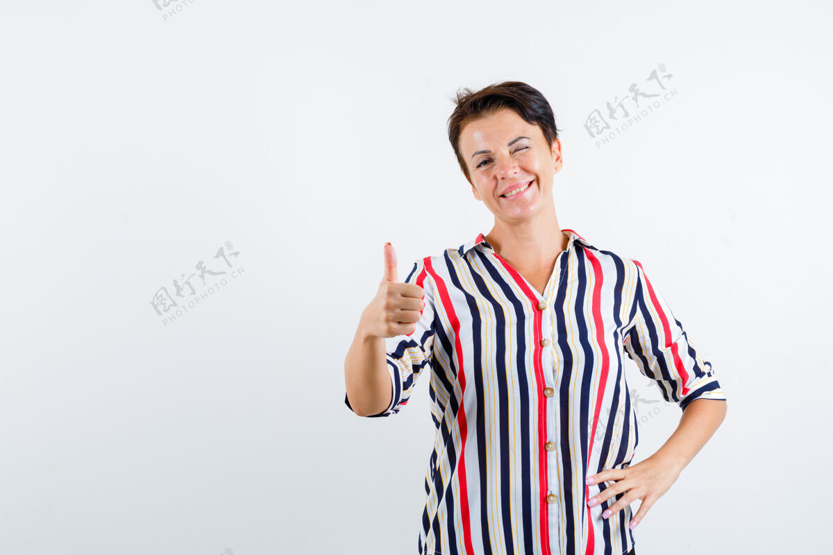 眨眼成熟的女人竖起大拇指 眨眼 手放在腰上 穿着条纹衬衫 看起来很自信前视图拇指人优雅