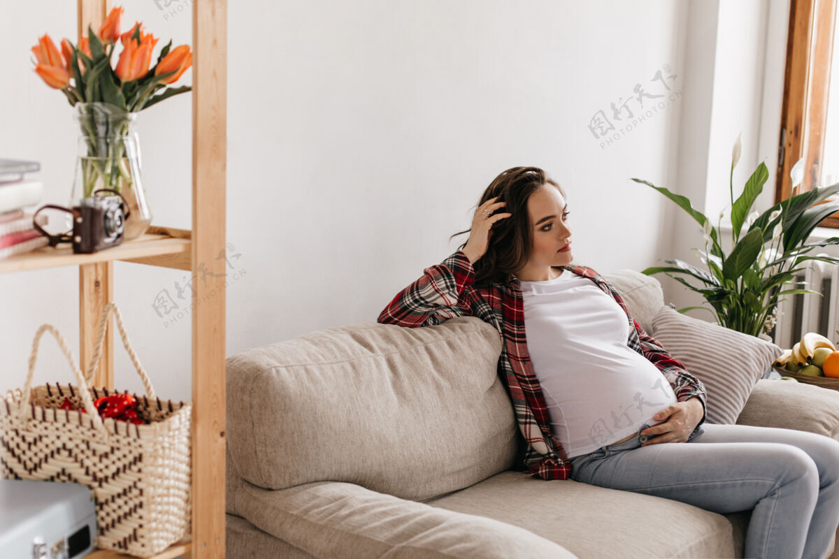 美丽卷曲的黑发孕妇穿着牛仔裤和格子衬衫躺在柔软的沙发上沙发腹部感觉