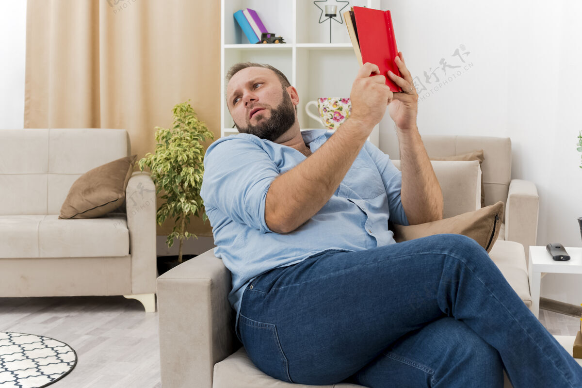 扶手椅未出租的成年斯拉夫男子坐在扶手椅上拿着书 看着客厅里的一面斯拉夫人男人壁板