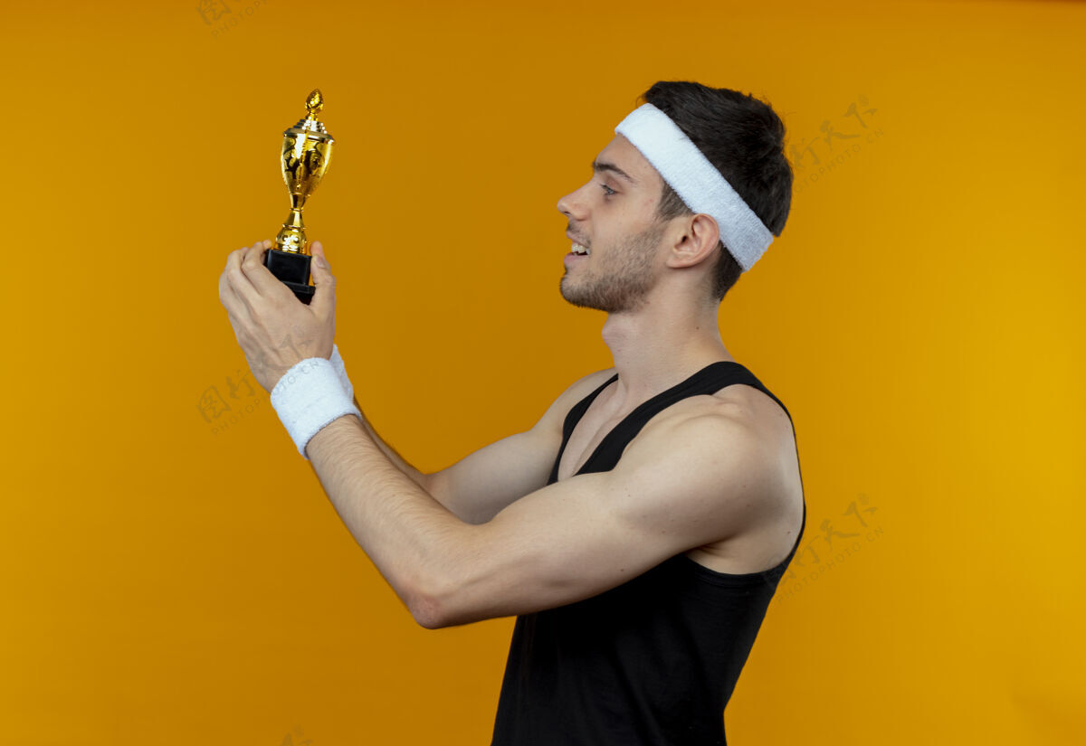 立场戴着头巾的年轻运动型男子拿着他的奖杯 站在橙色背景下 看着我快乐而积极的微笑年轻持有积极