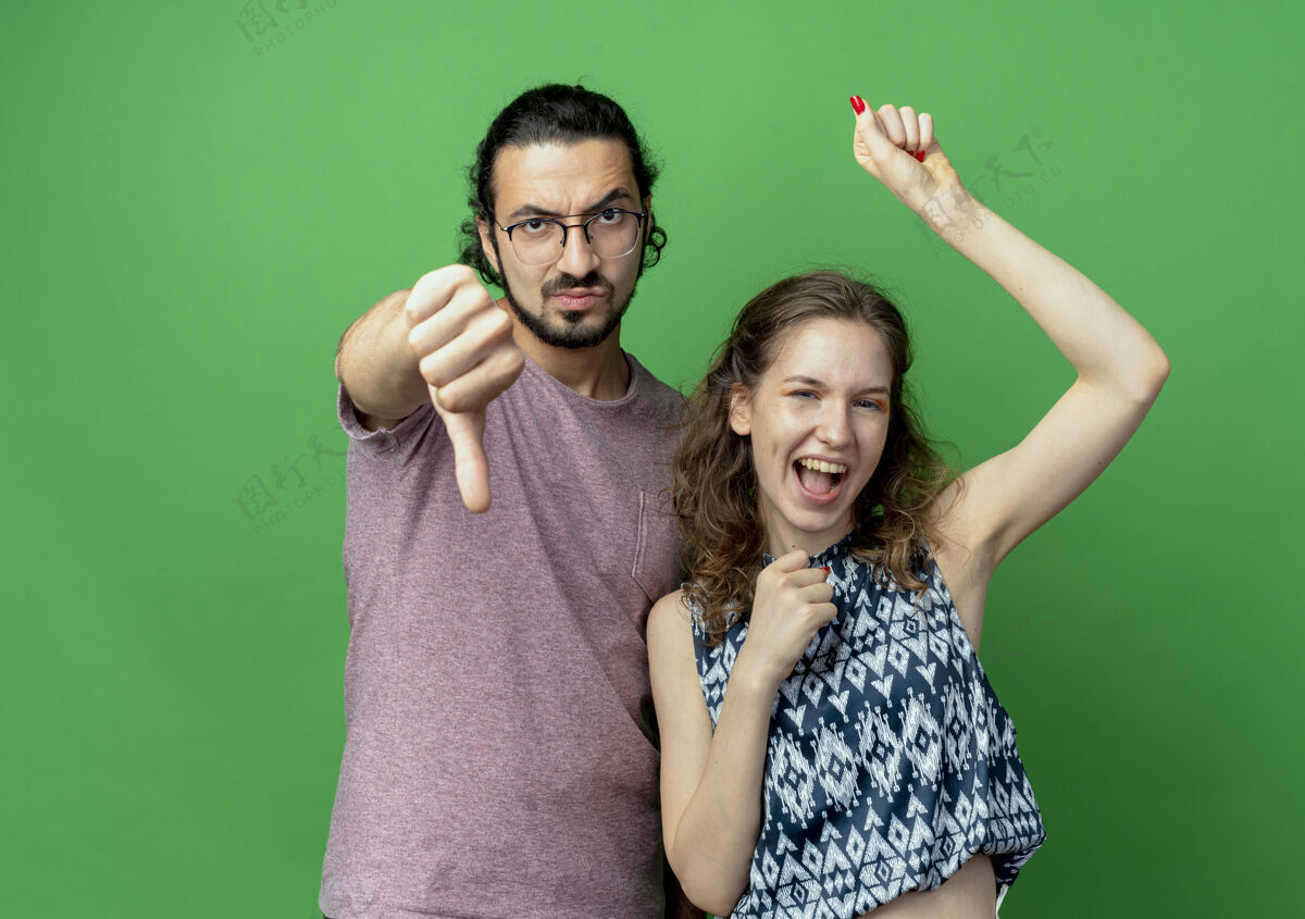 年轻这对年轻夫妇不高兴的男子在绿色背景下站在他快乐的女友旁边竖起大拇指站着不高兴一对