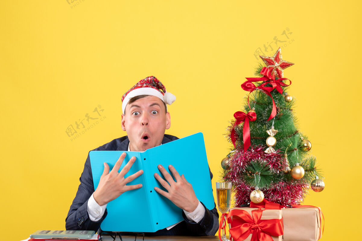 前面前视图惊奇的商人坐在圣诞树旁的桌子上 呈现黄色背景坐着成人微笑