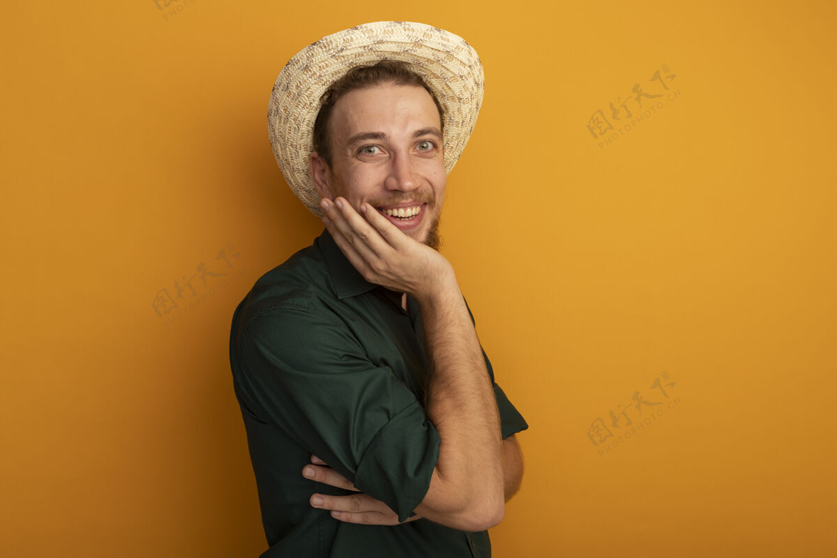 站着面带微笑的金发帅哥戴着沙滩帽把手放在隔离在橙色墙上的脸上感觉衣服帽子