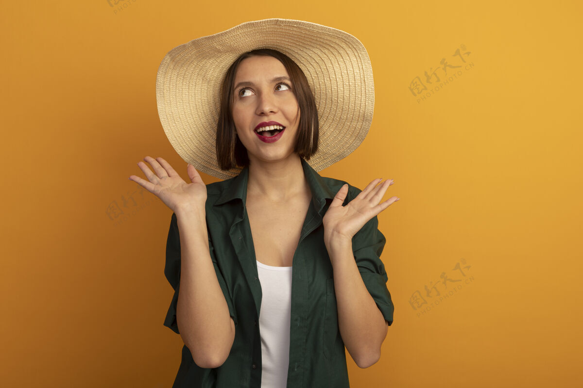 人戴着沙滩帽的快乐美女在橙色的墙上张开双手姿势举行开放