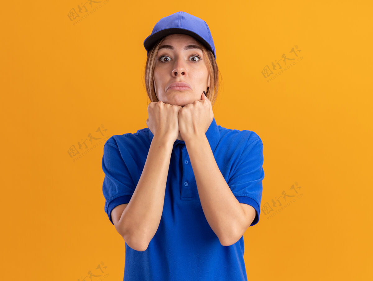人焦急的年轻漂亮的女送货员穿着制服 把拳头放在下巴上 看着隔离在橙色墙上的前面感情脸焦虑