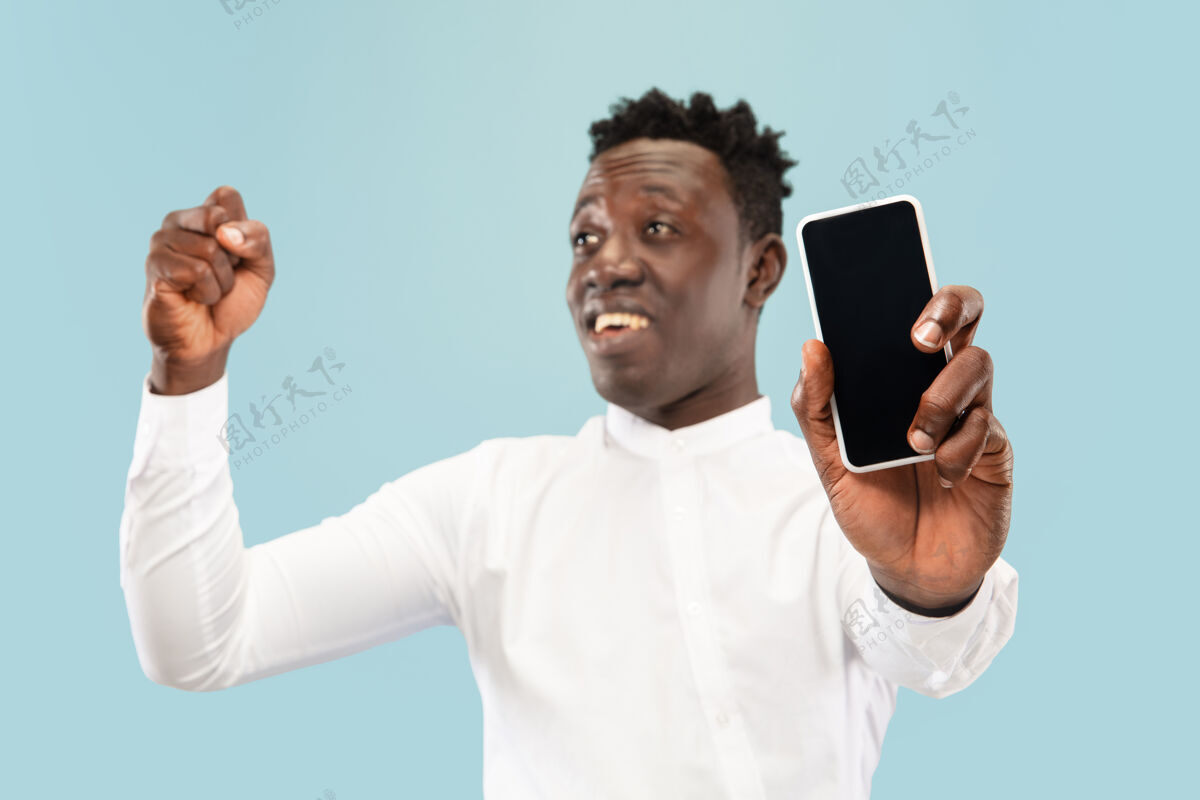 非洲裔年轻的非洲裔美国男子与智能手机在蓝色工作室背景上单独合影 面部表情美丽的男性半身肖像人类情感的概念 面部表情学生魅力有趣
