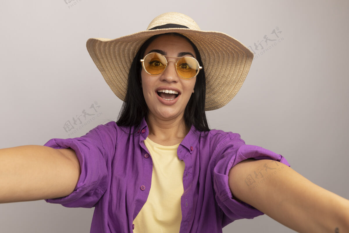 假装兴奋的黑发美女戴着沙滩帽 戴着太阳眼镜 假装站在前面 独自在白墙上自拍采取衣服人