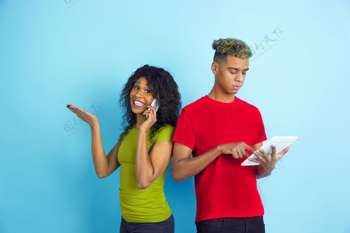 笑在平板电脑上工作 在电话上交谈年轻情绪化的非裔美国人男女在蓝色背景上穿着五颜六色的衣服美丽的情侣人类情感的概念 面部表情 关系 广告情侣头发美国