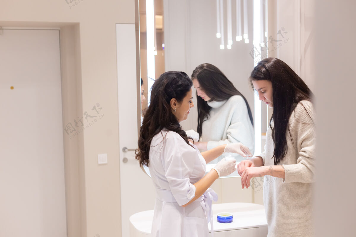 专业两个女人 一个美容师医生和一个客户 站在镜子前 为了咨询 医生把面霜涂在女人的手上 轻轻地按摩健康放松治疗
