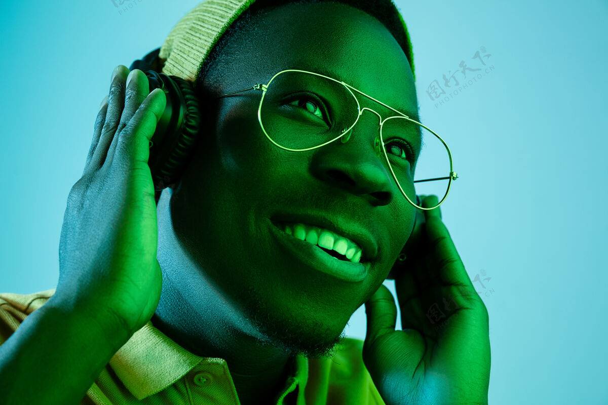 美国年轻帅气的快乐惊喜的时髦男人在霓虹灯下用耳机听音乐迪斯科舞厅 夜总会 嘻哈风格 积极的情绪 面部表情 舞蹈概念男性帽子娱乐