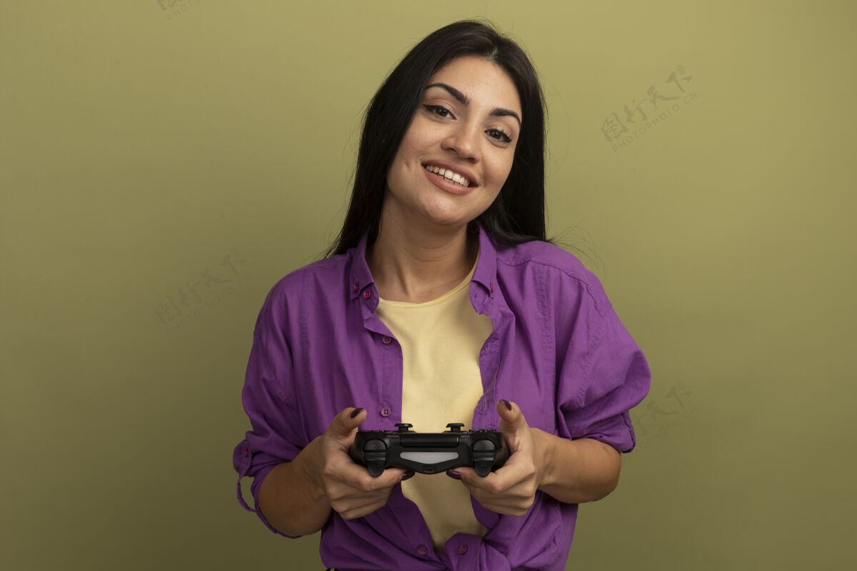 脸笑容可掬的黑发女子把游戏控制器孤立在橄榄绿的墙上控制器姿势衣服