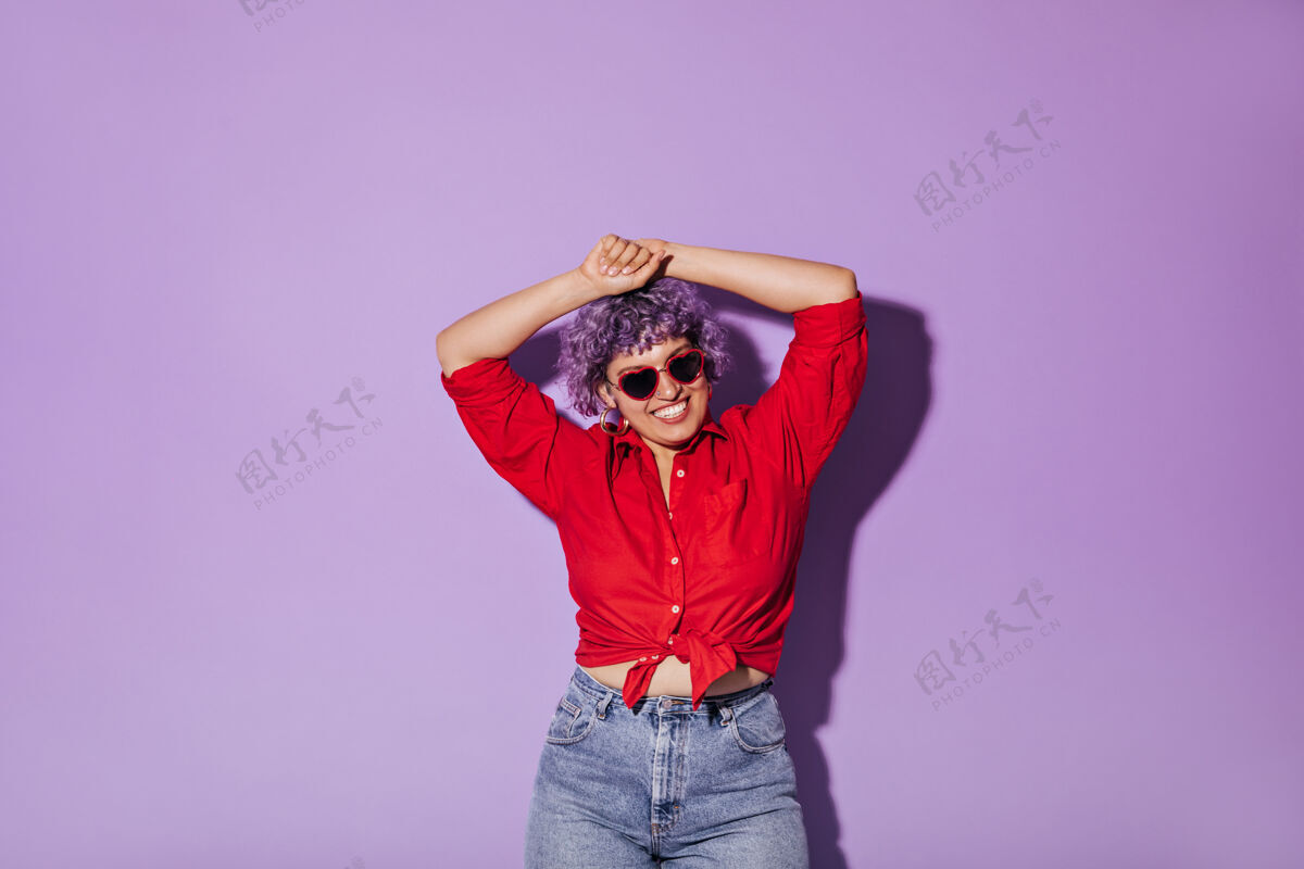 红衬衫身着红衫 长袖 戴着心形眼镜的快乐的现代年轻女子在紫色的舞台上摆姿势模特牛仔裤牛仔