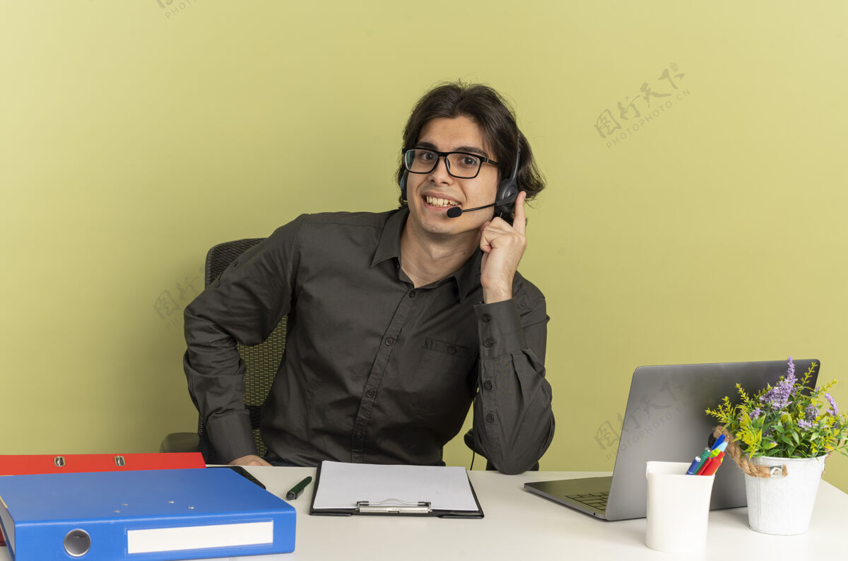 年轻戴着眼镜 面带微笑的年轻上班族坐在办公桌旁 手里拿着办公工具 手里拿着笔记本电脑 手放在绿色背景上隔离的耳机上 还有复印空间微笑眼镜办公室