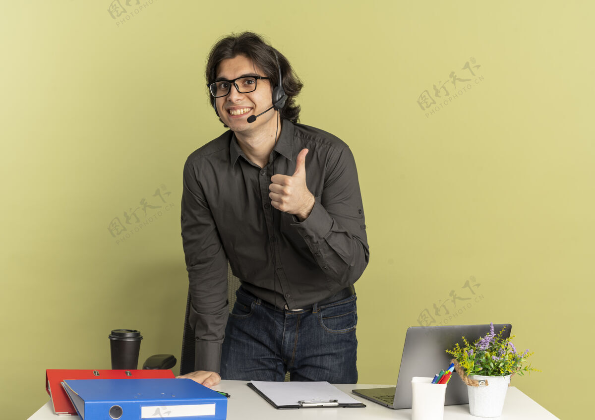 笔记本电脑戴着耳机 戴着眼镜 面带微笑的年轻上班族站在办公桌旁 拿着办公工具 用笔记本电脑 竖起大拇指 隔离在绿色背景上 留有复印空间拇指工人工具