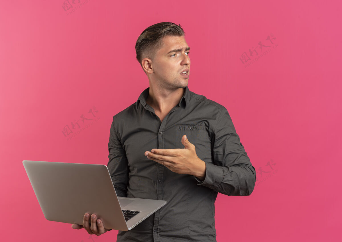 笔记本电脑年轻恼怒的金发帅哥拿着笔记本电脑 看着粉色背景上有空白的一面英俊恼怒尖