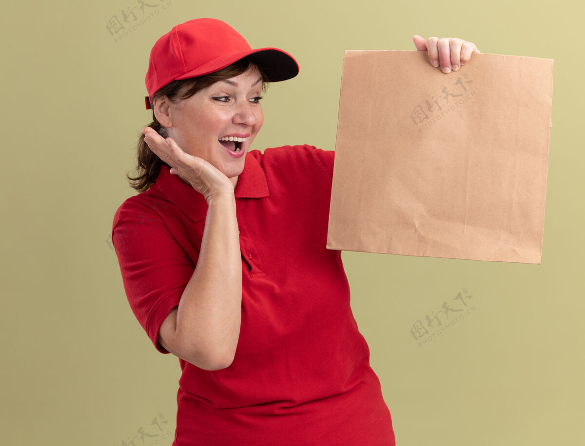 中年中年送货员身穿红色制服 戴着帽子 手里拿着纸包 站在绿色的墙边 高兴而兴奋地看着它包装制服送货