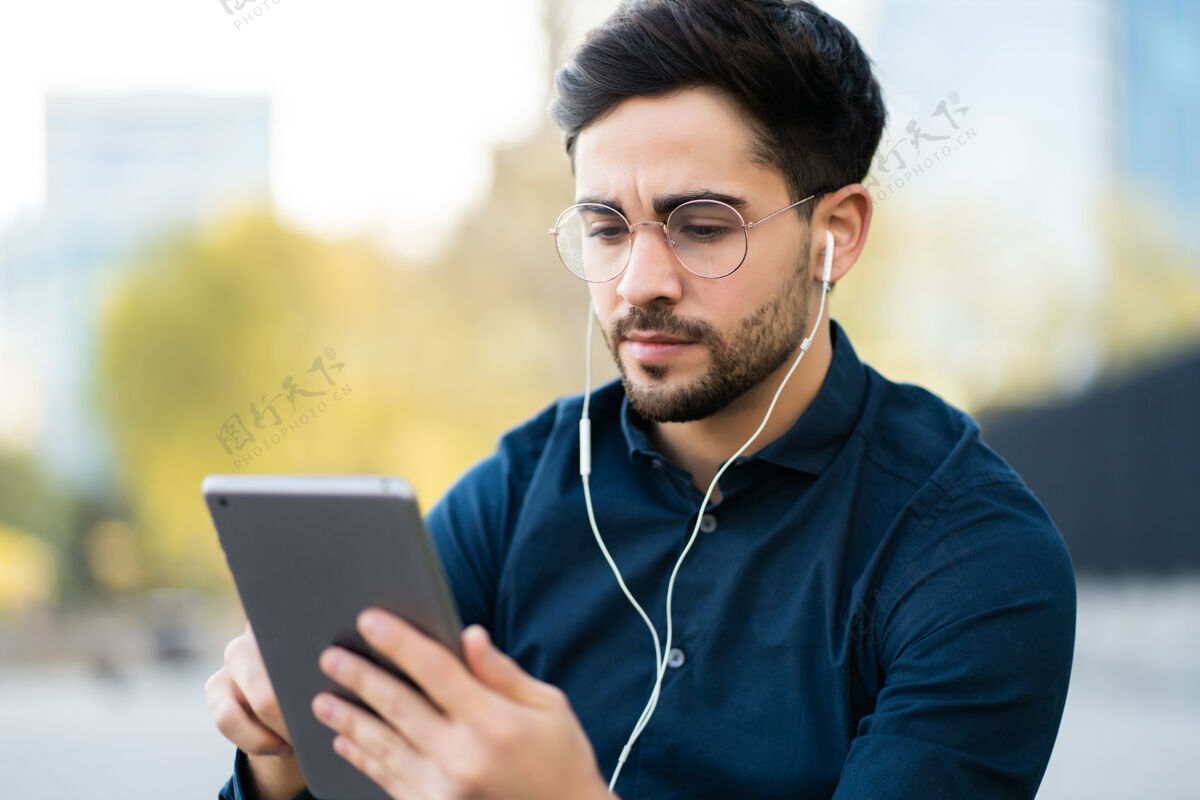 数字坐在户外长凳上使用数码平板电脑的年轻人肖像城市概念城市耳机男性