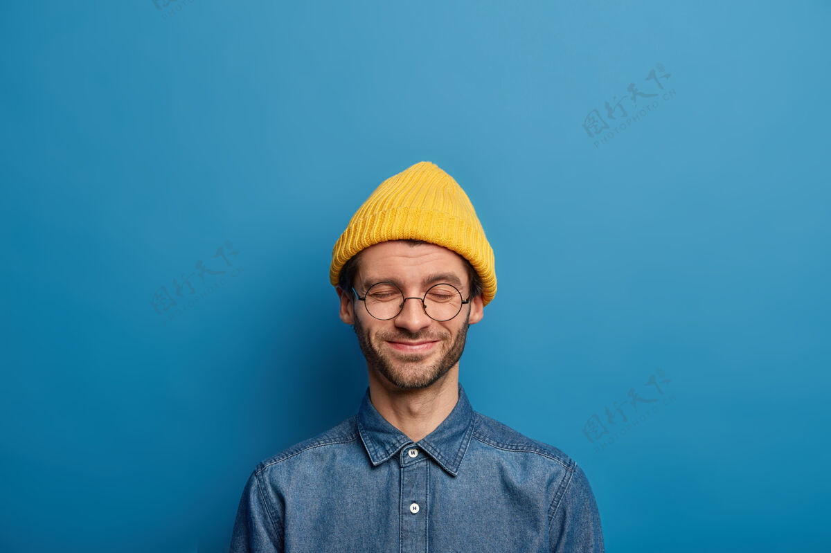 头饰乐观的高加索男人闭着眼睛 开心地笑着 戴着黄色的头饰和牛仔衬衫学生请沉思