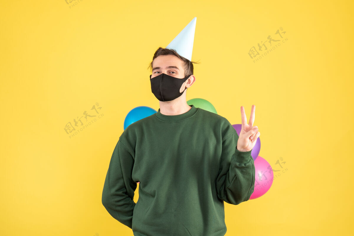 人物正面图：戴着派对帽的年轻人把气球藏在背后站在黄色背景上肖像胡子背面