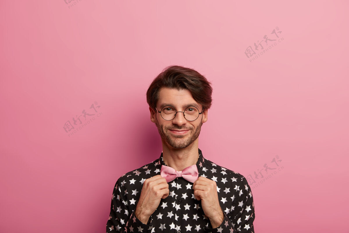 学生满意的未刮胡子的时髦男人手拉粉色领结 高兴地看着自己 穿着约会或会议的衣服 戴着光学圆眼镜请男性优雅