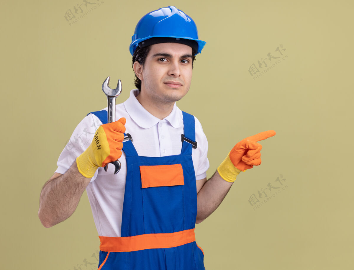 扳手年轻的建筑工人穿着建筑制服 戴着安全帽 戴着橡胶手套 拿着扳手 严肃地看着前面 用食指指着站在绿色墙上的一边建筑工人壁板安全