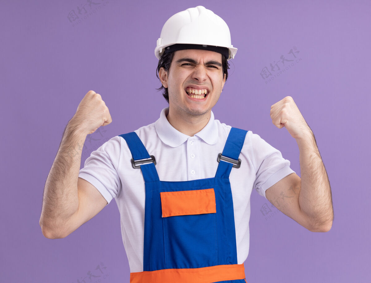 安全年轻的建筑工人穿着建筑制服 戴着安全帽 高兴而兴奋地握紧拳头 为他成功地站在紫色的墙上而欢呼欢喜兴奋男人